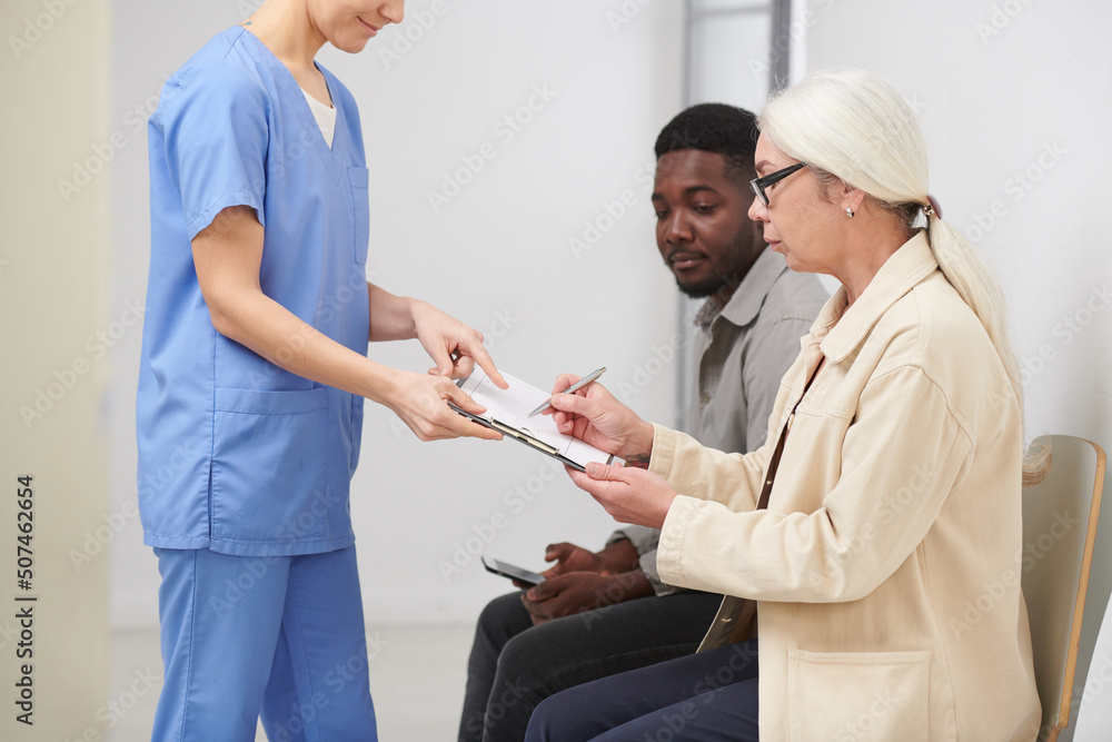 Obraz premium Side view shot of stylish senior woman sitting in queue in hospital signing important medical document papers