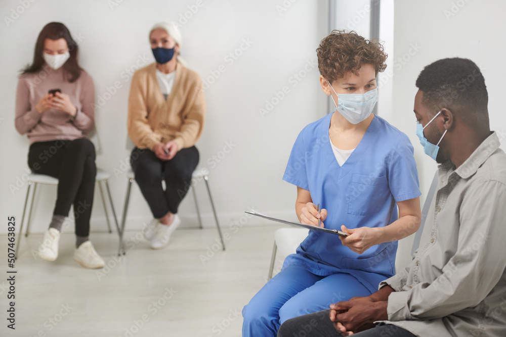 Obraz premium Horizontal shot of unrecognizable medical worker wearing mask sitting on chair in corridor next to patient asking him questions and making notes