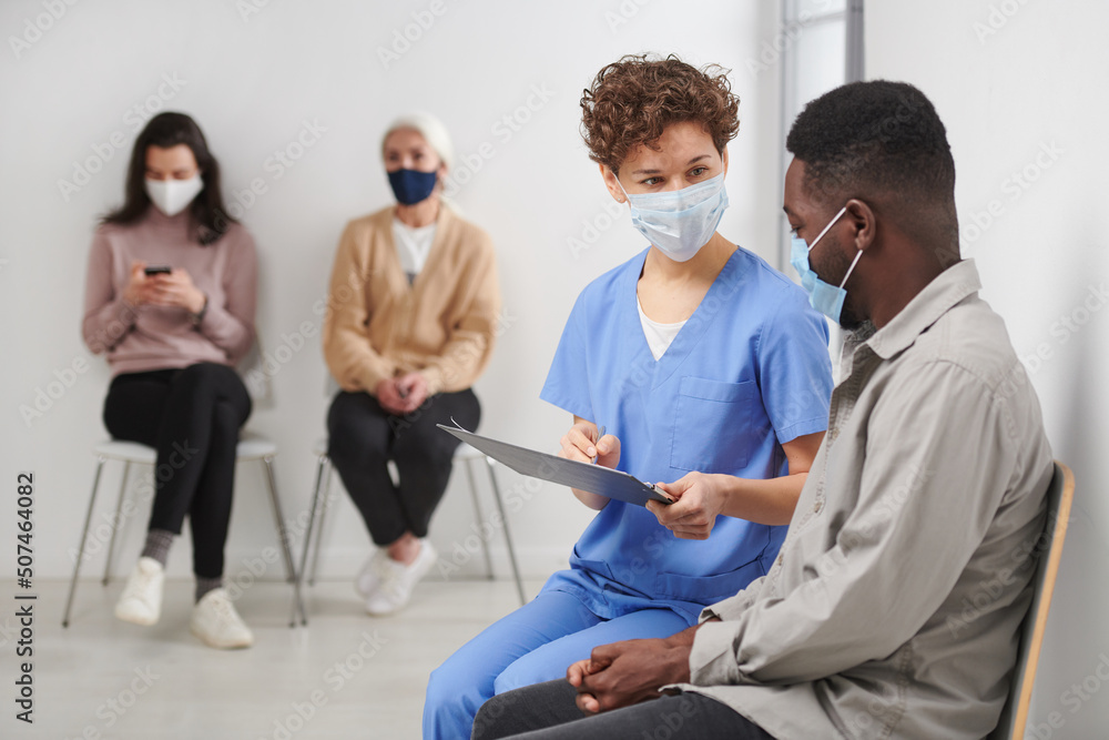 Horizontal shot of unrecognizable female medical worker wearing protective mask sitting on chair in corridor next to patient talking to him about test results Horizontal shot of unrecognizable female medical worker wearing protective mask sitting on chair in corridor next to patient talking to him about test results