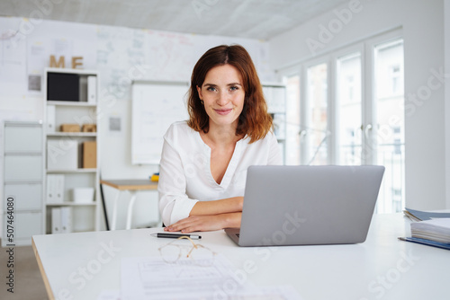 Fotografering young woman sits in the office in front of her laptop and looks into the camera