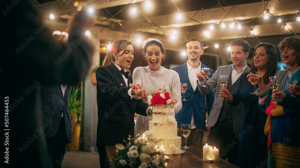 Beautiful Happy Lesbian Couple Celebrate Wedding at an Evening Reception Party with Diverse Multiethnic Friends. Queer Married Couple Standing at a Dinner Table, Kiss and Cut Wedding Cake. Beautiful Happy Lesbian Couple Celebrate Wedding at an Evening Reception Party with Diverse Multiethnic Friends. Queer Married Couple Standing at a Dinner Table, Kiss and Cut Wedding Cake.