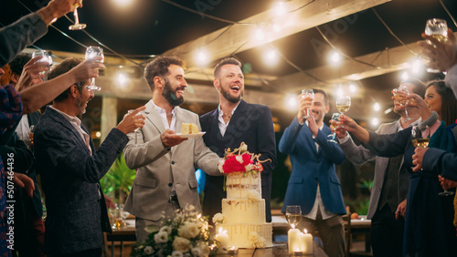 Handsome Happy Gay Couple Celebrate Wedding at an Evening Reception Party with Diverse Multiethnic Friends. Queer Married Couple Standing at a Dinner Table, Kiss and Cut Wedding Cake.