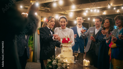 Beautiful Happy Lesbian Couple Celebrate Wedding at an Evening Reception Party with Diverse Multiethnic Friends. Queer Married Couple Standing at a Dinner Table, Kiss and Cut Wedding Cake.