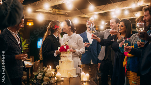 Beautiful Happy Lesbian Couple Celebrate Wedding at an Evening Reception Party with Diverse Multiethnic Friends. Queer Married Couple Standing at a Dinner Table, Kiss and Cut Wedding Cake.