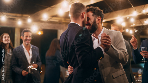 Handsome Happy Gay Couple Celebrate Wedding at an Evening Reception Party with Diverse Multiethnic Friends. Queer Newlyweds Dancing and Kissing at a Restaurant Venue. LGBTQ Relationship Goals.