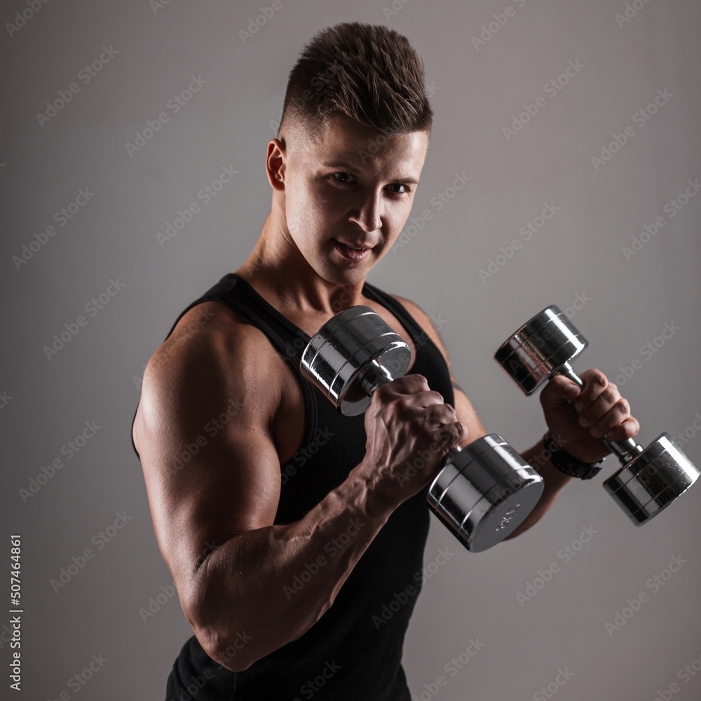 Strong handsome young man athlete model with a bare muscular torso workout with metal dumbbells on a dark background Strong handsome young man athlete model with a bare muscular torso workout with metal dumbbells on a dark background