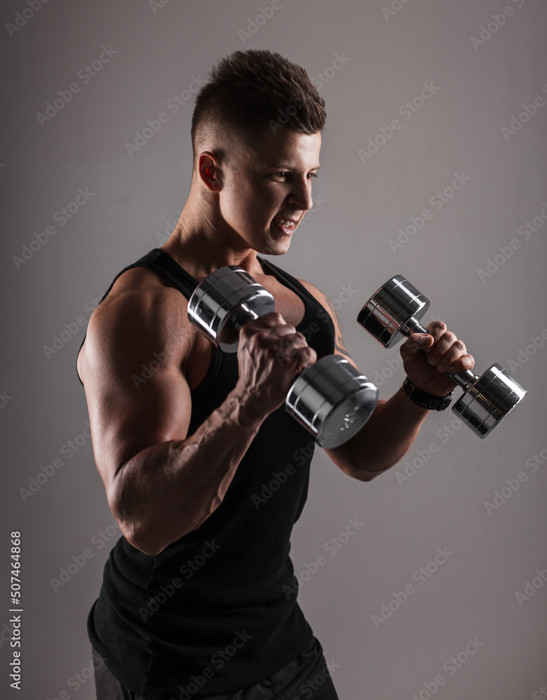 Powerful young handsome athletic male bodybuilder with a muscular body in black tank top is training hard with dumbbells on a dark background in the studio Powerful young handsome athletic male bodybuilder with a muscular body in black tank top is training hard with dumbbells on a dark background in the studio