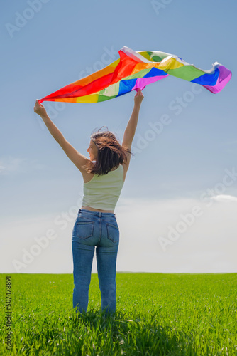 A woman waves a rainbow LGBT flag standing with back on a meadow against the background of the horizon and the sky looks towards Equal human rights in relation to the LGBT community