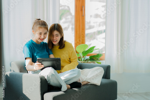 Two Asian women sit on gray sofas holding tablets while watching movies and listening to music on social media in their home living room.