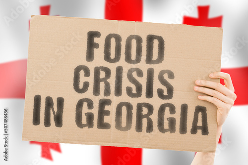 The phrase " Food crisis in Georgia " is on a banner in men's hands with a blurred Saudi Georgian flag in the background. Crisis. Finance. Life. Nutrition. Bread. Disaster. Collapse. Social issue