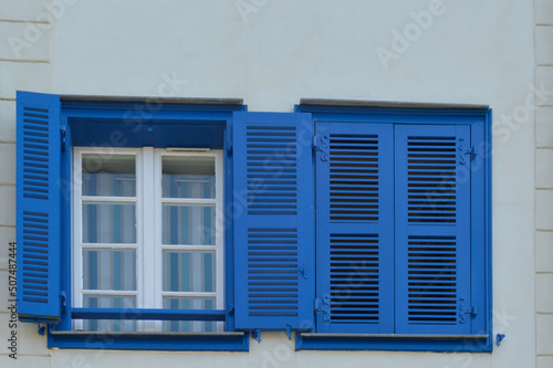 Une fenêtre blanche avec un volet bleu ouvert et une fenêtre aux volets fermés