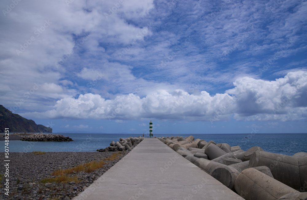 A landscape with a striped lighthouse at the end of the pier under the blue sky with white clouds on a sunny day. Skyline with a lighthouse, concrete tetrapods, Funchal, Madeira, Atlantic Ocean.