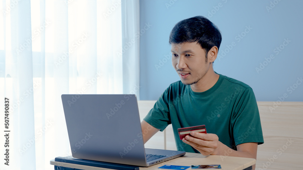 Handsome Asian boy, wearing a green t-shirt, is picking up his credit card, chooses to pay by credit card, while shopping online,shopping online. Handsome Asian boy, wearing a green t-shirt, is picking up his credit card, chooses to pay by credit card, while shopping online,shopping online.