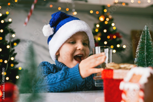 Boy toddler in a blue Santa hat drinking filtered water from a glass in the kitchen. Holidays, health concept.