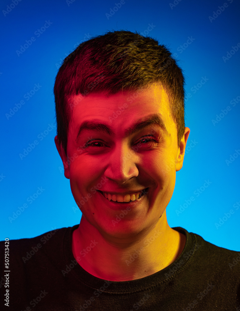 Obraz premium Handsome emotional man's portrait on blue studio background in orange neon, multicoloured. Very fun and stupid laughing, narrowing our eyes, we look at the camera and show our teeth. Concept of huma