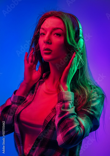 Portraits of young emotional woman on multicoloured background in neon lights. Listening to music with headphones with pleasure and enjoyment. Looks with interest, adjusts glasses. Flyer Poster Banner