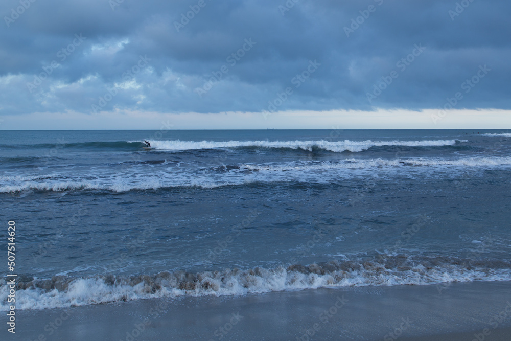 Lone surfer riding a wave under a stormy sky with tanker ship on distant horizon Lone surfer riding a wave under a stormy sky with tanker ship on distant horizon