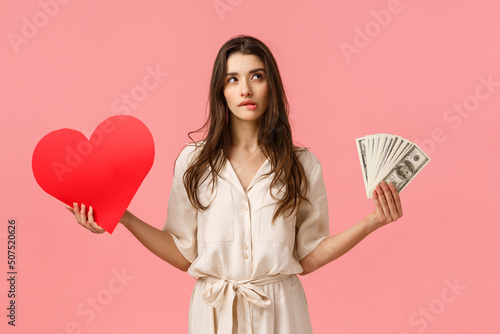 Romance, wealth and modern lifestyle concept. Alluring good-looking brunette female in dress, holding cash, money and love heart, biting lip look upper right corner unsure, making decision photo