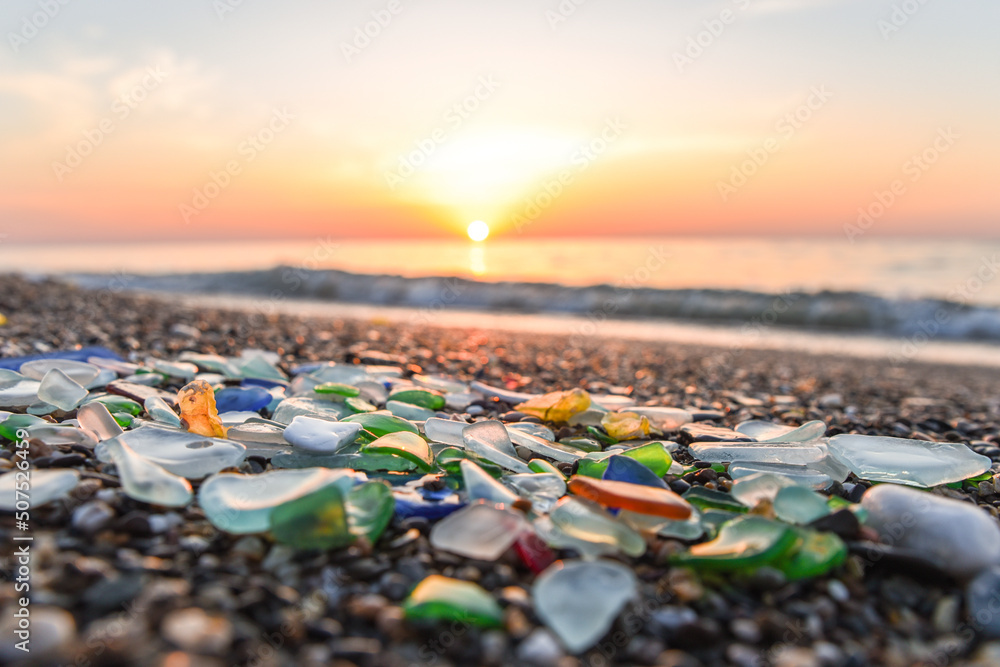 Colored sea glass with beach pebbles and shells in the mediterranean coast and in the background sea and waves with sunset and sky, sandy beach with coast glass, beach glass. - obrazy, fototapety, plakaty 