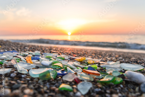 Colored sea glass with beach pebbles and shells in the mediterranean coast and in the background sea and waves with sunset and sky, sandy beach with coast glass, beach glass.