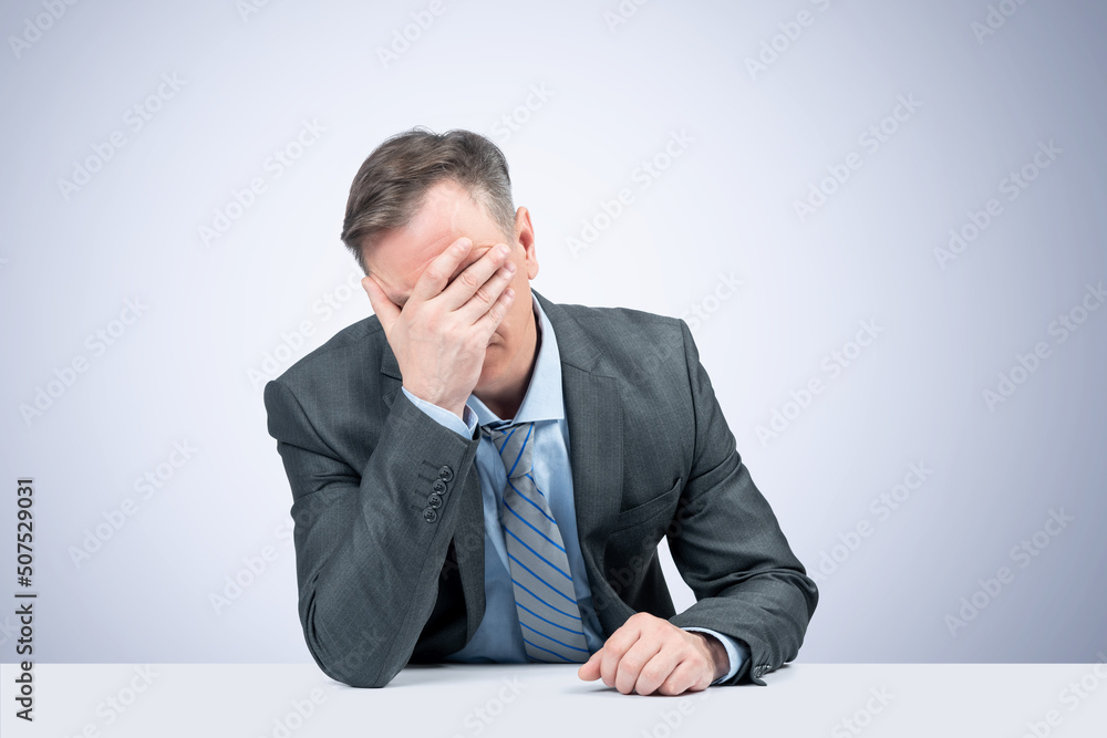 A man in a dark jacket, shirt and tie at the table makes a facepalm, on light blue background. Epic fail emotion. A man in a dark jacket, shirt and tie at the table makes a facepalm, on light blue background. Epic fail emotion.