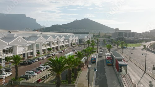 Fly along large shopping centre on Victoria and Alfred Waterfront. Traffic on wide street surrounded by palm trees. Cape Town, South Africa