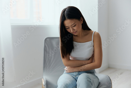 Pregnancy lost. Abortion high. Tormented suffering tanned beautiful young Asian woman hold hands on stomach at home interior living room. Injuries Poor health Illness concept. Cool offer Banner