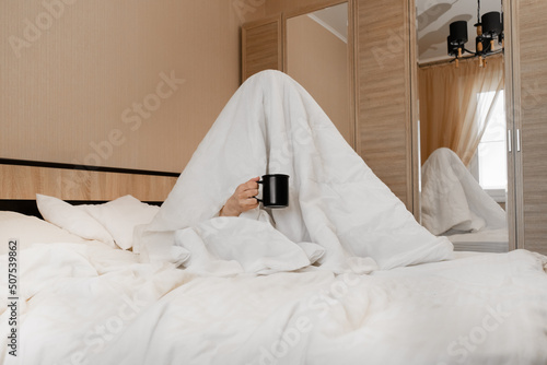 A person under a blanket with a cup of coffee can not cheer up, does not want to get out of bed and hides from the world. A hard morning. Lack of sleep.