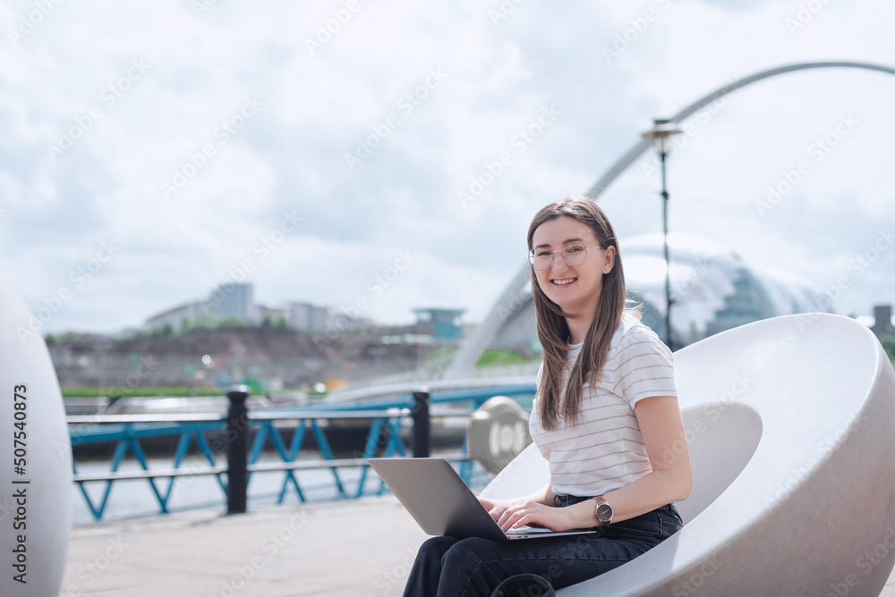 A young woman works at her laptop, sitting on a modern bench on the Newcastle waterfront, against the backdrop of Sage Gateshead and Gateshead Millennium. North East England.