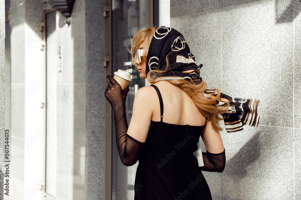 Naklejka premium Back view of young woman with long fair hair wearing black dress, long mesh gloves, silk printed kerchief, sunglasses.