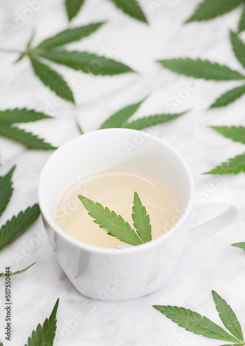 white mug with marijuana infusion with green leaf inside on marble table with leaves. top view.