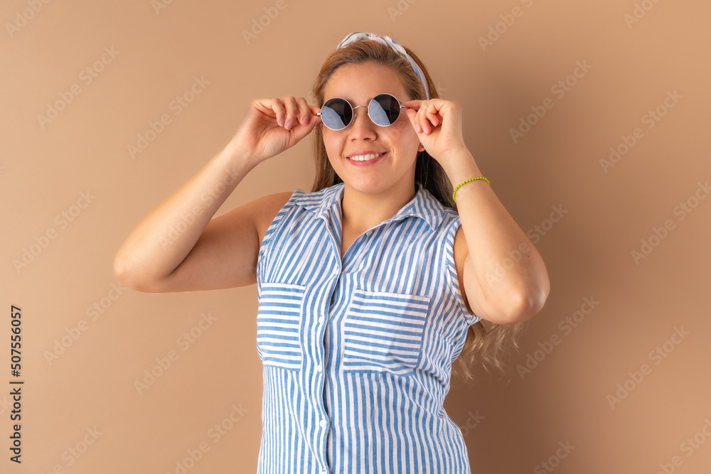 Beautiful attractive young woman posing happily with trendy sunglasses, in striped blue dress. Isolated over brown background.