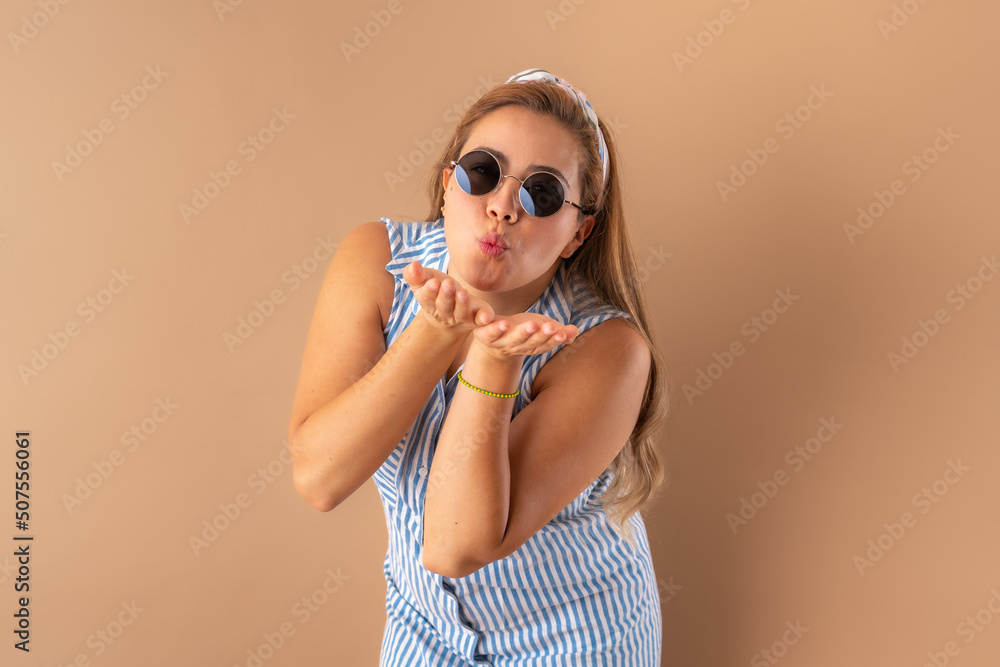 Beautiful attractive young woman posing happily with trendy sunglasses and sending an air kiss, in striped blue dress. Isolated over brown background. Beautiful attractive young woman posing happily with trendy sunglasses and sending an air kiss, in striped blue dress. Isolated over brown background.