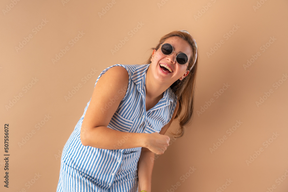 Beautiful attractive young woman posing happily with trendy sunglasses, in striped blue dress. Isolated over brown background.