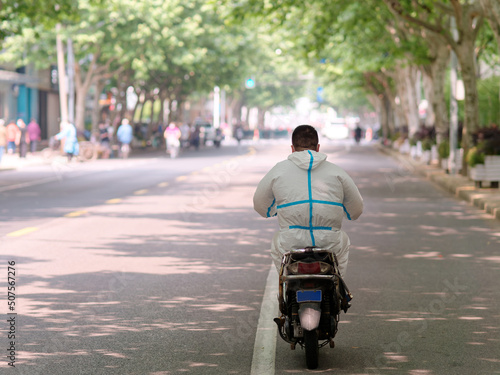 Rear view of man wearing hazmat suit driving electrical bicycle on road in sunny day, Shanghai city lockdown for two months, few people can go outside because of quarantine policy. photo