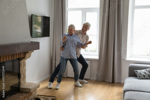 Active retirement. Happy energetic older age married couple dancing at modern living room with electric chimney feel healthy having fun. Funny joyful grandparents relax dance indoors by favorite music © fizkes