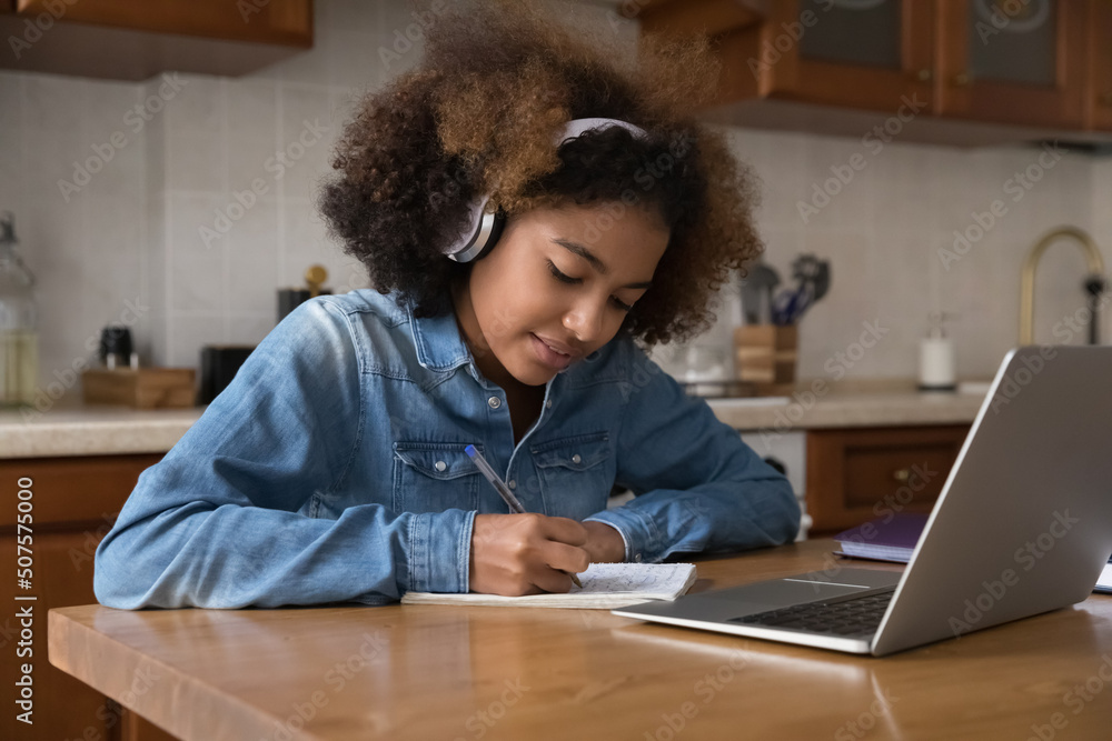 Serious busy high school student teenage girl in wireless headphones studying at home, writing notes, sitting at kitchen table, using laptop, watching video class, learning webinar Serious busy high school student teenage girl in wireless headphones studying at home, writing notes, sitting at kitchen table, using laptop, watching video class, learning webinar