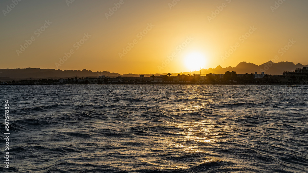 Sunset over the Red Sea. The sun is low over a picturesque mountain range. The sky is colored orange. Ripples and highlights on the water. Egypt. Safaga
