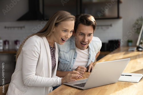 Happy surprised millennial couple using laptop, staring at screen, getting good awesome news, mortgage approval, winning prize, unexpected money, income, benefit, excited with discount, sale