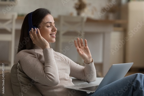Calm happy pretty girl in big wireless headphones listening to ambient tranquil music from online playlist, enjoying tunes for stress relief, meditation, relaxation, using laptop with closed eyes