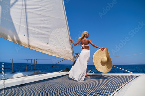 Leinwand Poster Beautiful young blond woman in bikini standing on catamaran at sunny summer day