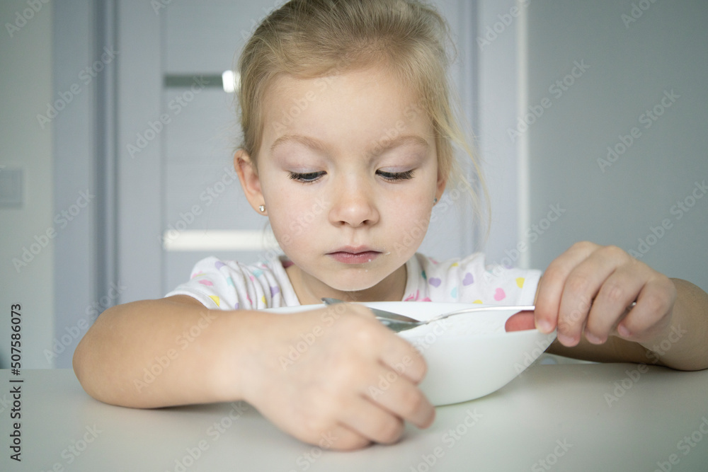 Child girl blonde eats with a spoon from a white plate at a white table. Child girl blonde eats with a spoon from a white plate at a white table.