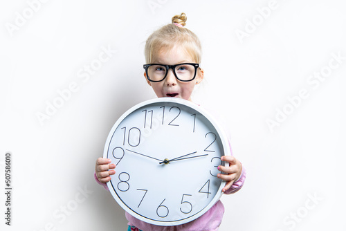Surprised child girl in glasses holding a large wall clock on a light background. photo
