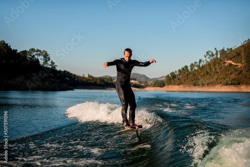 man in wetsuit energetically balancing on river water on a foil wakeboard on beautiful landscape background.