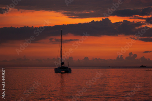 Majestic saturated sea landscape - colorful sunset in ocean with golden sky, dark clouds, reflections in calm water, orange sunbeams, silhouette floated yacht. Marine landscape on indonesian island.