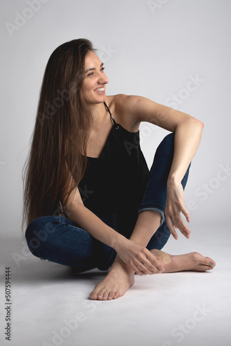 Beauty, fashion and make-up concept. Portrait of happy, beautiful, slim, tall and sexy looking woman with long dark hair wearing black blouse and blue jeans sitting on white studio background floor
