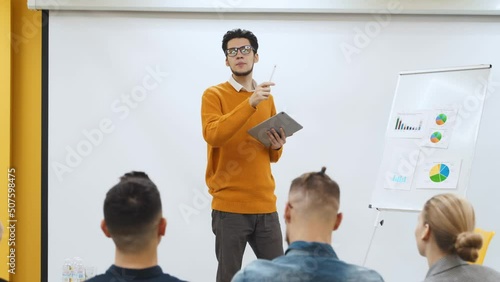 Businessman coach trainer presents report, shows diagrams on flip chart in conference hall. Audience raises hands for answer on presentation in office. Teacher leading master class to audience. photo