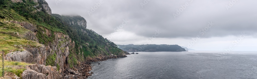 Panoramic shot of green karst cliffs of Cabo Antzoriz and grey, rippling waters of Bay of Biscay on cloudy, rainy summer morning. Lequeitio, Biscay, Basque Country