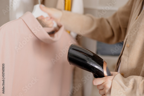 Young woman steaming her clothes at home, close up view. Female using electric steamer, ironing clothes on hanger. Modern technologies.