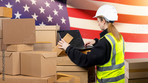 Woman with boxes and laptop. USA customs check concept. Customs officer woman. Girl with computer in front American flag. Lady in yellow vest with her back to camera. Customs clearance goods from USA photo